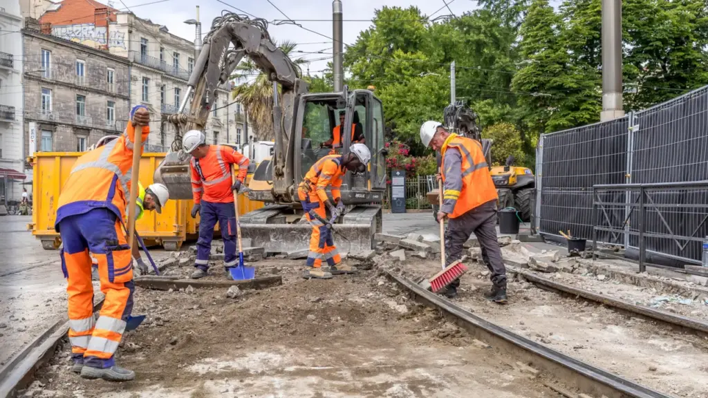 Des ouvriers en gilets orange travaillent sur des rails de tramway près d'une pelleteuse.