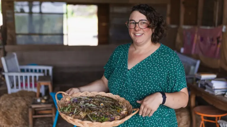 Une femme souriante aux cheveux courts tenant un plateau en osier rempli de plantes.
