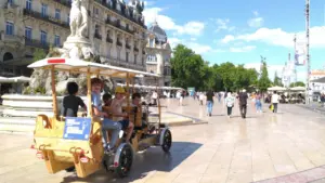 Un groupe d'enfants souriants monte un vélo-bus en bois sur la place de la Comédie.