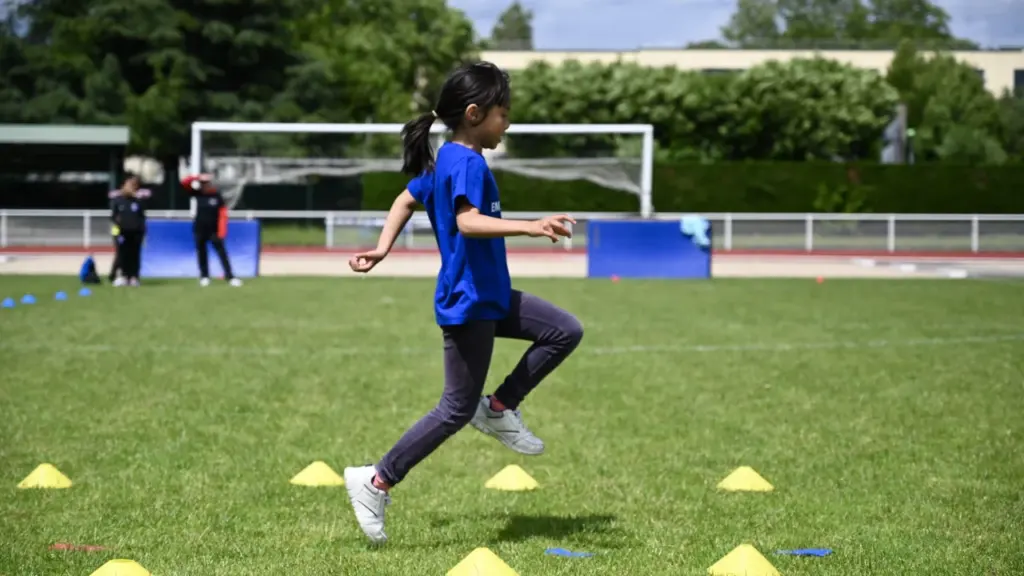Une jeune fille en t-shirt bleu court sur une pelouse parsemée de cônes jaunes.