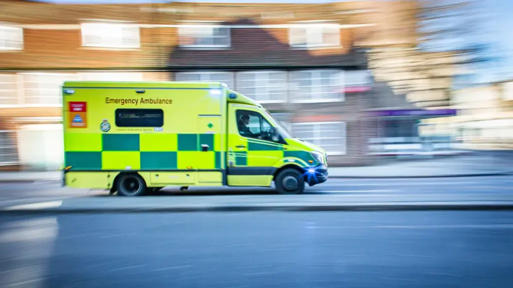 Une ambulance jaune et verte circulant rapidement dans une rue résidentielle en journée.