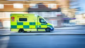 Une ambulance jaune et verte circulant rapidement dans une rue résidentielle en journée.