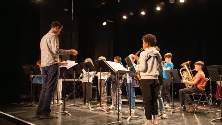 Un chef d'orchestre dirige un groupe d'élèves musiciens jouant divers instruments sur scène.