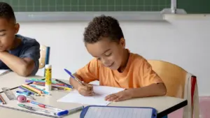 Un jeune garçon souriant écrit sur une feuille de papier dans une salle de classe.