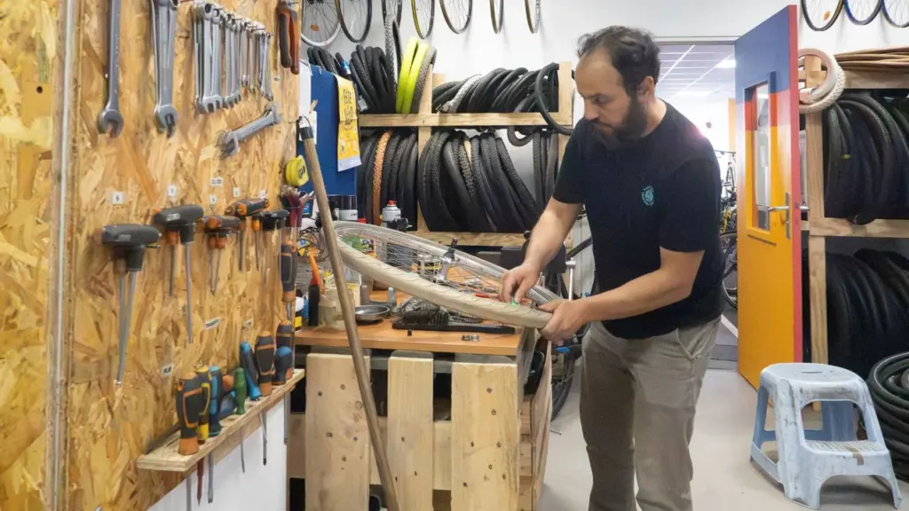 Un mécanicien barbu répare une roue de vélo dans un atelier rempli d'outils suspendus.
