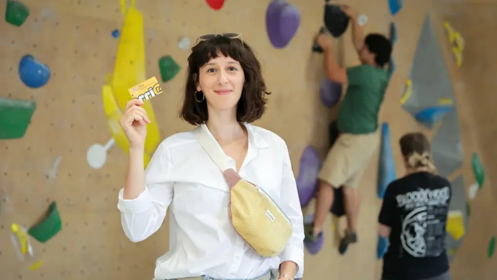 Une femme souriante tenant une carte devant un mur d'escalade avec des prises colorées.