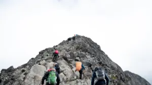 Un groupe de randonneurs équipés gravissant un sommet montagneux rocheux sous un ciel couvert.