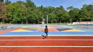 Un jeune garçon court sur une piste d'athlétisme rouge devant un terrain de basket coloré.