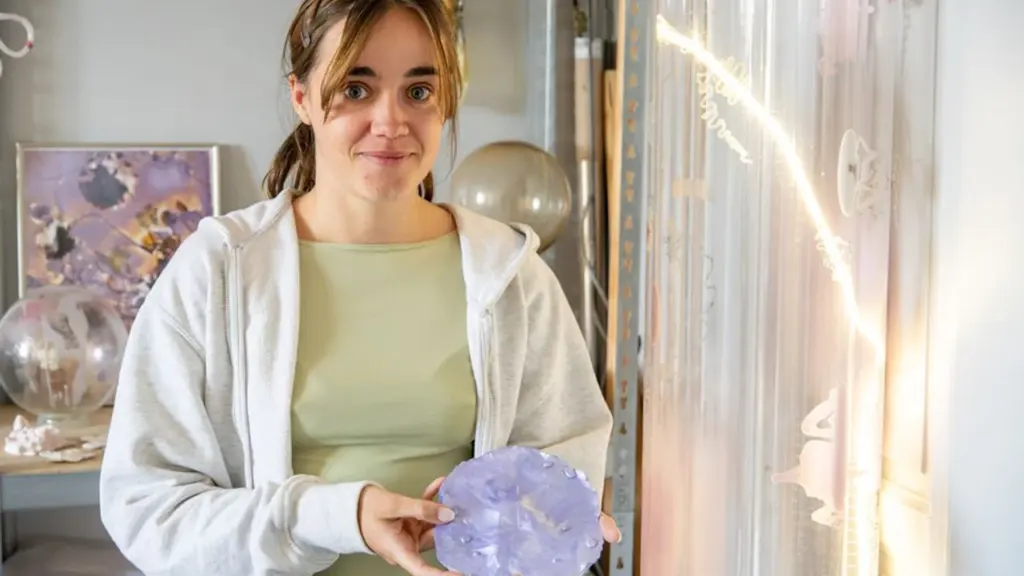 Une jeune femme aux cheveux châtains tient un objet circulaire en verre violet sculpté.